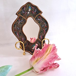 May include: A decorative mirror with a bronze-coloured frame, adorned with a floral and bird design in blue, pink, and green. The mirror is supported by a gold-coloured stand. A pink tulip is in the foreground. The mirror's frame is approximately 20 cm in height.