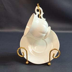 Op de afbeelding: Een witte theekop en schotel set gepresenteerd op een goudkleurige metalen standaard. De theekop is gekanteld, waardoor de binnenkant zichtbaar is, en de schotel ligt eronder. De standaard heeft een decoratief ontwerp.