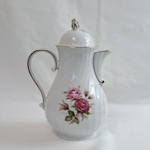 Puede incluir: Cafetera de porcelana blanca con ribete dorado y un diseño floral con rosas rosas. La cafetera tiene una tapa con un pomo en forma de rosa.