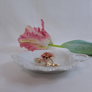 Könnte beinhalten: Eine weiße, blattförmige Schale aus Keramik mit einer rosa und grünen Blume und zwei goldenen Broschen mit rosa Steinen.