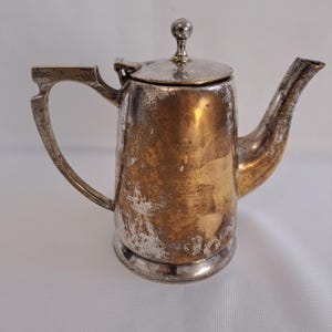 Könnte beinhalten: Vintage silberbeschichtete Teekanne mit angelaufenem Aussehen. Die kleine Teekanne hat einen gebogenen Ausguss, einen Griff mit dekorativem Design und einen Deckel mit einem kleinen Knopf. Das Metall hat Gold- und Silbertöne.