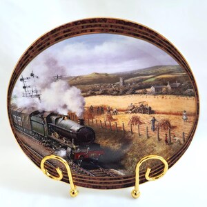 Op de afbeelding: Decoratieve plaat met een gedetailleerd schilderij van een stoomtrein op rails, met rook, en een landelijke oogstscène. De plaat heeft een bruine en gouden rand en wordt gepresenteerd op een goudkleurige standaard.