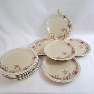 Op de afbeelding: Een set beige keramische borden met een bloemenpatroon. Elk bord heeft een roze bloem en bruin blad. De borden zijn gerangschikt op een wit oppervlak, sommige gestapeld en andere tentoongesteld op een gouden bordenstandaard.