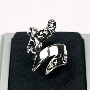 Può includere: Un anello d'argento con un designo dettagliato e ornato che presenta un serpente stilizzato con una coda arricciata. L'anello è esposto su uno sfondo di velluto nero.