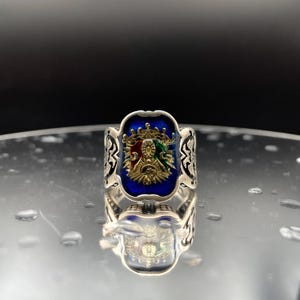 Pode incluir: Um anel de prata com um design de esmalte azul que apresenta um brasão dourado com detalhes em vermelho, verde e dourado. O anel tem detalhes intrincados em prata ao redor das bordas.