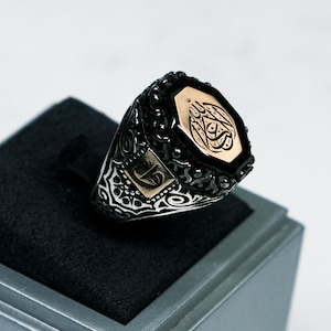 以下が含まれることがあります： 八角形の黒オニキスがゴールドトーンのベゼルにセットされた、黒と銀のリング。石には複雑なアラビア語の書道が施されています。