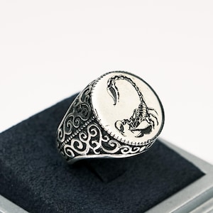 以下が含まれることがあります： 蠍の彫刻が施された楕円形のトップを持つシルバーリング。リングのバンドにも渦巻き模様が細かく彫刻されています。