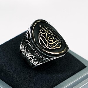 Può includere: Un anello d'argento con un designo ornato e un centro ovale nero con calligrafia araba dorata.