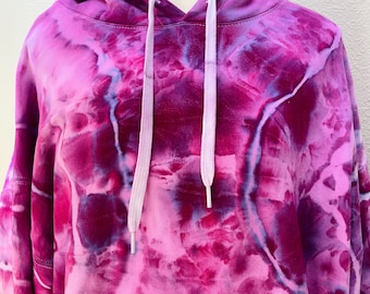 May include: A tie-dye hoodie in shades of pink and purple, with white drawstrings. The hoodie features a front pocket and a hood. The fabric has a soft, cloud-like pattern. The garment is displayed on a mannequin.