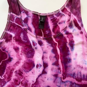 May include: A sleeveless tank top with a tie-dye pattern in shades of pink and purple. The fabric has a marbled effect, with streaks of lighter pink and white. The neckline and armholes are trimmed with a contrasting dark pink stitching.
