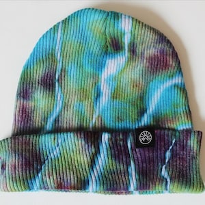 May include: A tie-dye beanie in shades of blue, green, and purple. The ribbed knit hat features a folded brim and a small black tag with a white mountain logo. The hat is a stylish accessory.