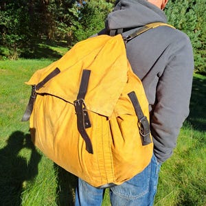 Könnte beinhalten: Ein großer senfgelber Rucksack aus Segeltuch mit einem umklappbaren Deckel, der mit drei dunkelbraunen Lederriemen und Metallschnallen gesichert ist. Die Tasche verfügt über verstellbare Schultergurte und ein geräumiges Hauptfach.