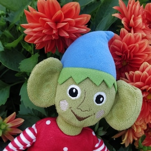 Könnte beinhalten: Eine Plüschpuppe mit hellgrünem Gesicht, großen Ohren und einer blauen Zipfelmütze. Die Puppe hat aufgestickte schwarze Augen, ein kleines Lächeln und rosa Wangen. Sie trägt ein rotes Oberteil mit weißen Punkten und rot-weiß gestreiften Ärmeln.
