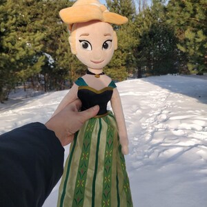 Op de afbeelding: Een pluche pop van een personage uit de animatiefilm Frozen. De pop heeft een blonde knot, een zwart en blauwgroen lijfje en een groen en goud gestreepte rok. De pop wordt vastgehouden tegen een besneeuwde achtergrond.