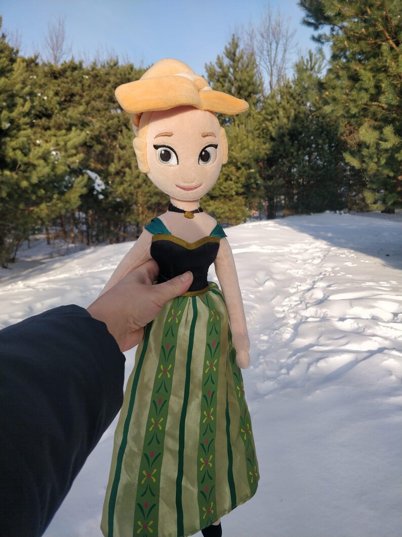 Op de afbeelding: Een pluche pop van een personage uit de animatiefilm Frozen. De pop heeft een blonde knot, een zwart en blauwgroen lijfje en een groen en goud gestreepte rok. De pop wordt vastgehouden tegen een besneeuwde achtergrond.