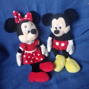 Puede incluir: Un conjunto de juguetes de peluche de Mickey Mouse y Minnie Mouse. Mickey lleva pantalones cortos rojos con lunares blancos y zapatos amarillos. Minnie lleva un vestido rojo con lunares blancos y zapatos rojos.