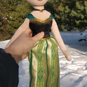 Op de afbeelding: Pluche pop van een personage uit de film Frozen, met een blonde knot en een zwarte en groene jurk. De pop heeft een zwarte choker en een goudkleurige ketting. De pop wordt vastgehouden tegen een besneeuwde achtergrond.
