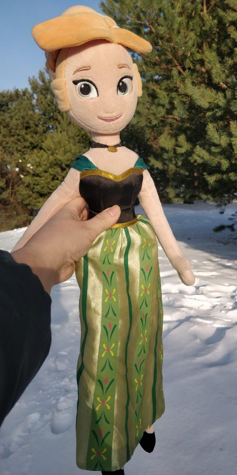 Op de afbeelding: Pluche pop van een personage uit de film Frozen, met een blonde knot en een zwarte en groene jurk. De pop heeft een zwarte choker en een goudkleurige ketting. De pop wordt vastgehouden tegen een besneeuwde achtergrond.