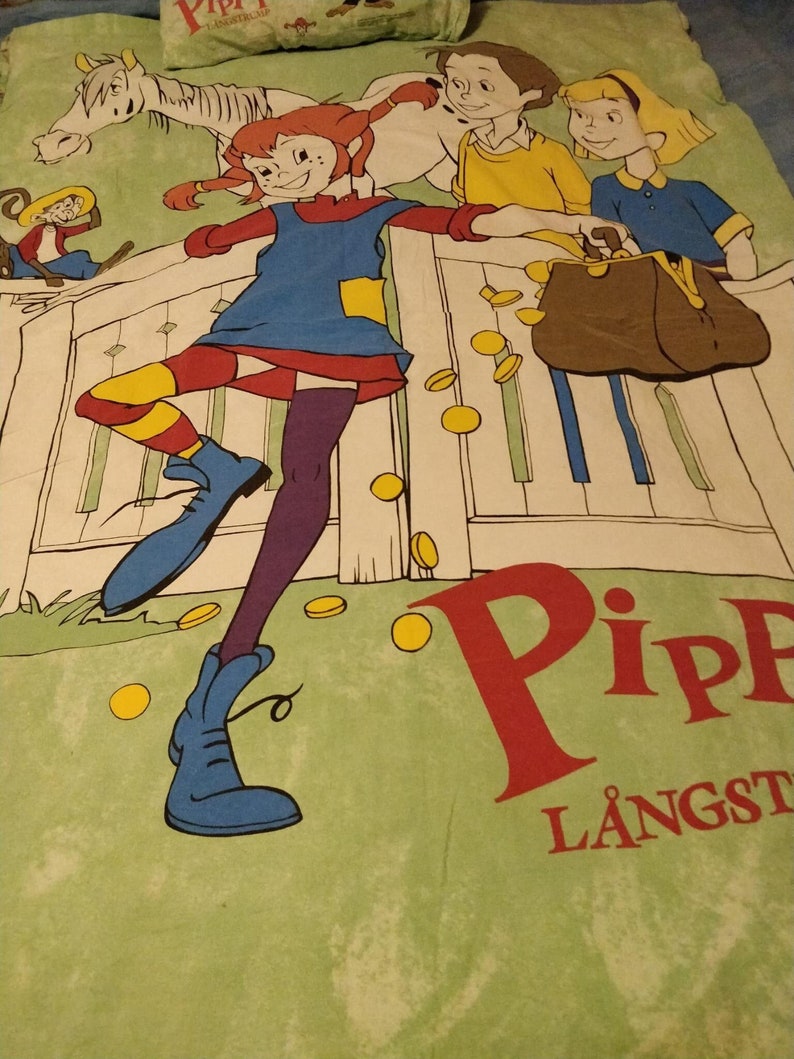 May include: A green and white bed sheet featuring a cartoon illustration of a girl with red hair and a blue dress, a boy with brown hair and a yellow shirt, and a girl with blonde hair and a blue shirt. The illustration includes a white horse, a brown purse, and gold coins. The text "Pippi L&aring;ngst" is printed in red and white on the sheet.