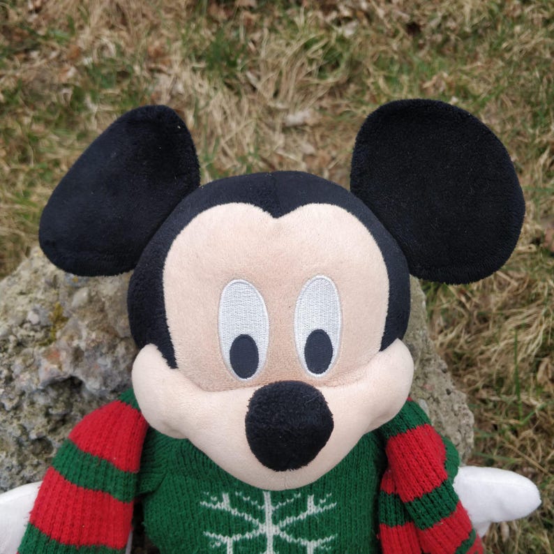 Peut inclure: Un Mickey Mouse en peluche portant un pull de No&euml;l vert et rouge. Le pull a un motif de flocon de neige blanc.