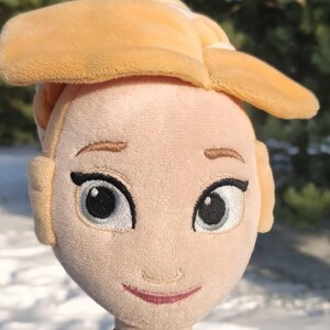 Op de afbeelding: Pluche pop van een personage met blond haar in een knot, geborduurde gezichtskenmerken en een zwarte choker met een gouden hanger. De pop draagt een groene en zwarte jurk. De achtergrond is een besneeuwd landschap met bomen.