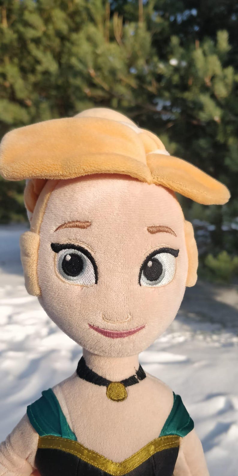 Op de afbeelding: Pluche pop van een personage met blond haar in een knot, geborduurde gezichtskenmerken en een zwarte choker met een gouden hanger. De pop draagt een groene en zwarte jurk. De achtergrond is een besneeuwd landschap met bomen.