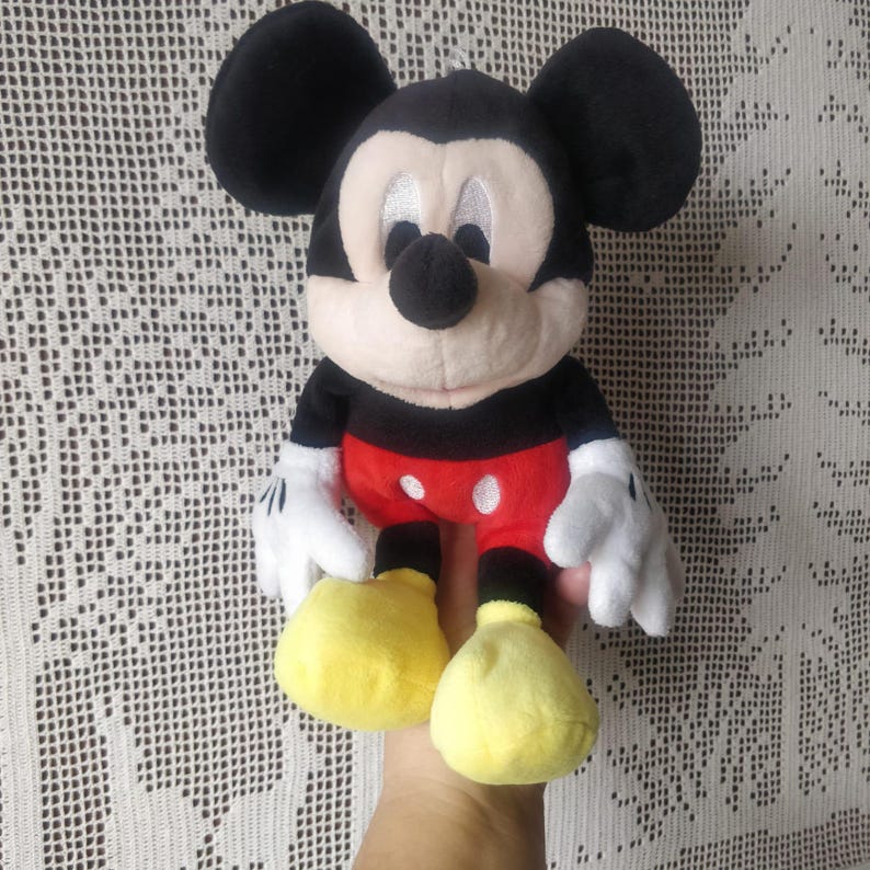 Puede incluir: Un juguete de peluche de Mickey Mouse con orejas negras, nariz negra y camisa roja con lunares blancos. El juguete est&aacute; sentado en una mano con zapatos amarillos.