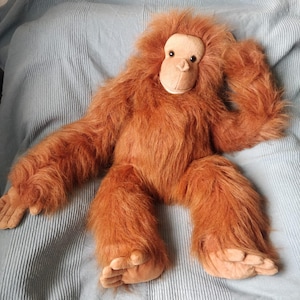 May include: A plush orangutan toy with long, shaggy, reddish-brown fur. The stuffed animal has a light tan face, hands, and feet, and is lying on a light blue, textured surface. The toy's arms and legs are outstretched.