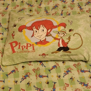 May include: A green and yellow checked fabric with a repeating pattern of a girl with red hair and a monkey. The fabric has the words "Pippi Langstrum" printed on it.