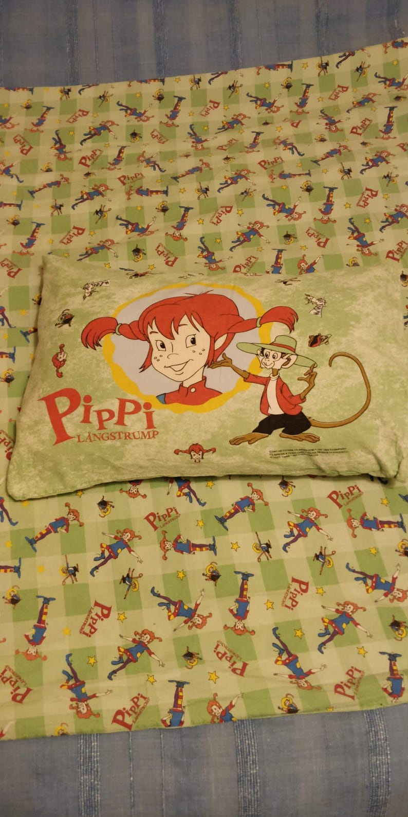 May include: A green and yellow checked fabric with a repeating pattern of a girl with red hair and a monkey. The fabric has the words "Pippi Langstrum" printed on it.