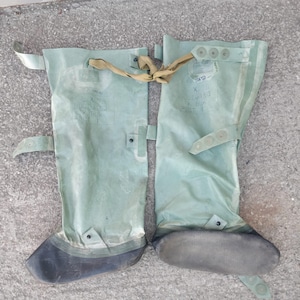 Op de afbeelding: Een paar lichtgroene waterdichte laarzen met zwarte zolen. De laarzen hebben riemen en gespen om ze vast te zetten. Er staat tekst op de laarzen. De laarzen zijn ontworpen om de benen en voeten tegen water te beschermen.