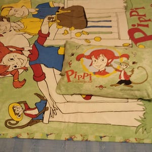 May include: A green and white bedspread featuring a cartoon image of Pippi Longstocking and her monkey, Mr Nilsson. The bedspread includes a pillow with a similar image of Pippi Longstocking and Mr Nilsson.