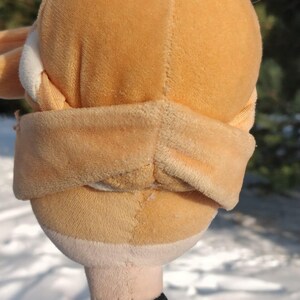 Op de afbeelding: Een pluche pop met een lichtoranje hoofd en een bijpassende blinddoek. De pop heeft een zwarte choker en een groen en zwart topje. De achtergrond is een besneeuwd landschap.