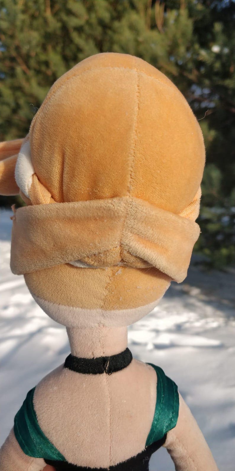 Op de afbeelding: Een pluche pop met een lichtoranje hoofd en een bijpassende blinddoek. De pop heeft een zwarte choker en een groen en zwart topje. De achtergrond is een besneeuwd landschap.
