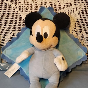 Könnte beinhalten: Ein Plüschtier von Mickey Mouse in einem hellblauen Strampler, mit schwarzen Ohren und einem lächelnden Gesicht. Das Spielzeug liegt auf einer hellblauen Decke mit gewelltem Rand. Der Hintergrund zeigt ein weißes Spitzendesign.