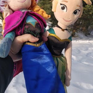 Op de afbeelding: Twee pluche poppen van animatiefiguren, een met een blauwe jurk en roze cape, de andere met een zwart lijfje en groene rok. Beide poppen hebben blond haar en lachen. De poppen worden vastgehouden tegen een besneeuwde achtergrond.