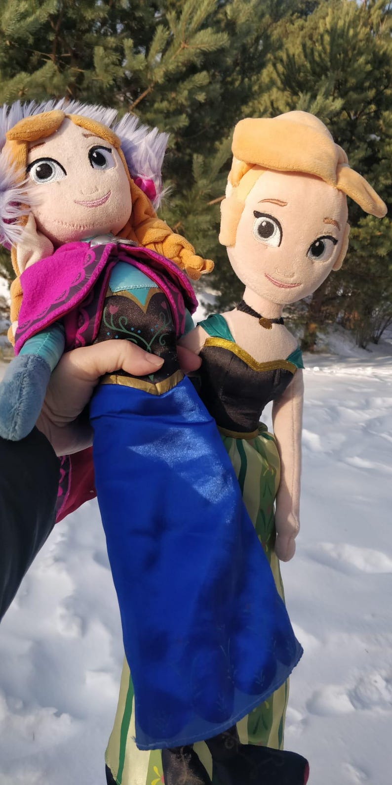 Op de afbeelding: Twee pluche poppen van animatiefiguren, een met een blauwe jurk en roze cape, de andere met een zwart lijfje en groene rok. Beide poppen hebben blond haar en lachen. De poppen worden vastgehouden tegen een besneeuwde achtergrond.