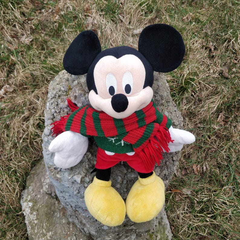 Peut inclure: Un jouet en peluche Mickey Mouse portant une &eacute;charpe ray&eacute;e rouge et verte. Le jouet est assis sur un rocher avec un fond herbeux vert.