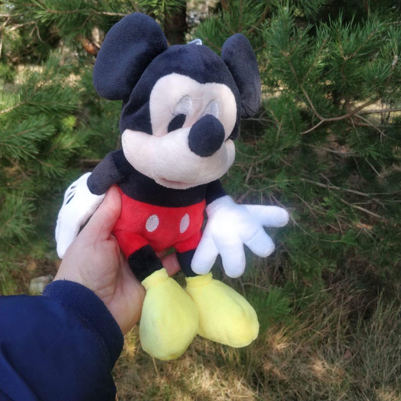 Puede incluir: Un juguete de peluche de Mickey Mouse con orejas negras, una cara negra, una camisa roja y blanca y zapatos amarillos. El juguete est&aacute; siendo sostenido por una mano.
