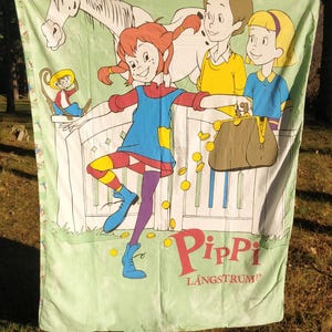 May include: A green and white fabric sheet featuring a cartoon image of Pippi Longstocking, a horse, a monkey, and two children. The text "Pippi Langstrum" is printed in red and white letters.