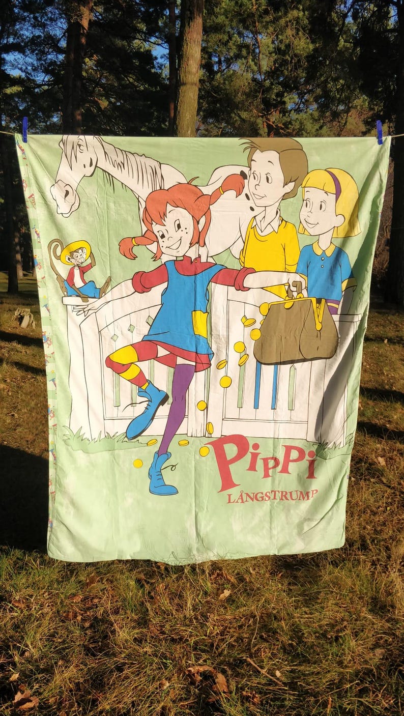May include: A green and white fabric sheet featuring a cartoon image of Pippi Longstocking, a horse, a monkey, and two children. The text "Pippi Langstrum" is printed in red and white letters.