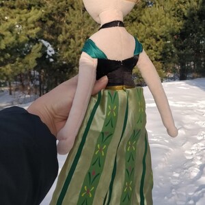 Op de afbeelding: Een pluche pop van een personage met blond haar in een knot. De pop draagt een zwarte top met groene accenten en een lange groene rok met een bloemenpatroon. De pop wordt vastgehouden tegen een besneeuwde achtergrond.