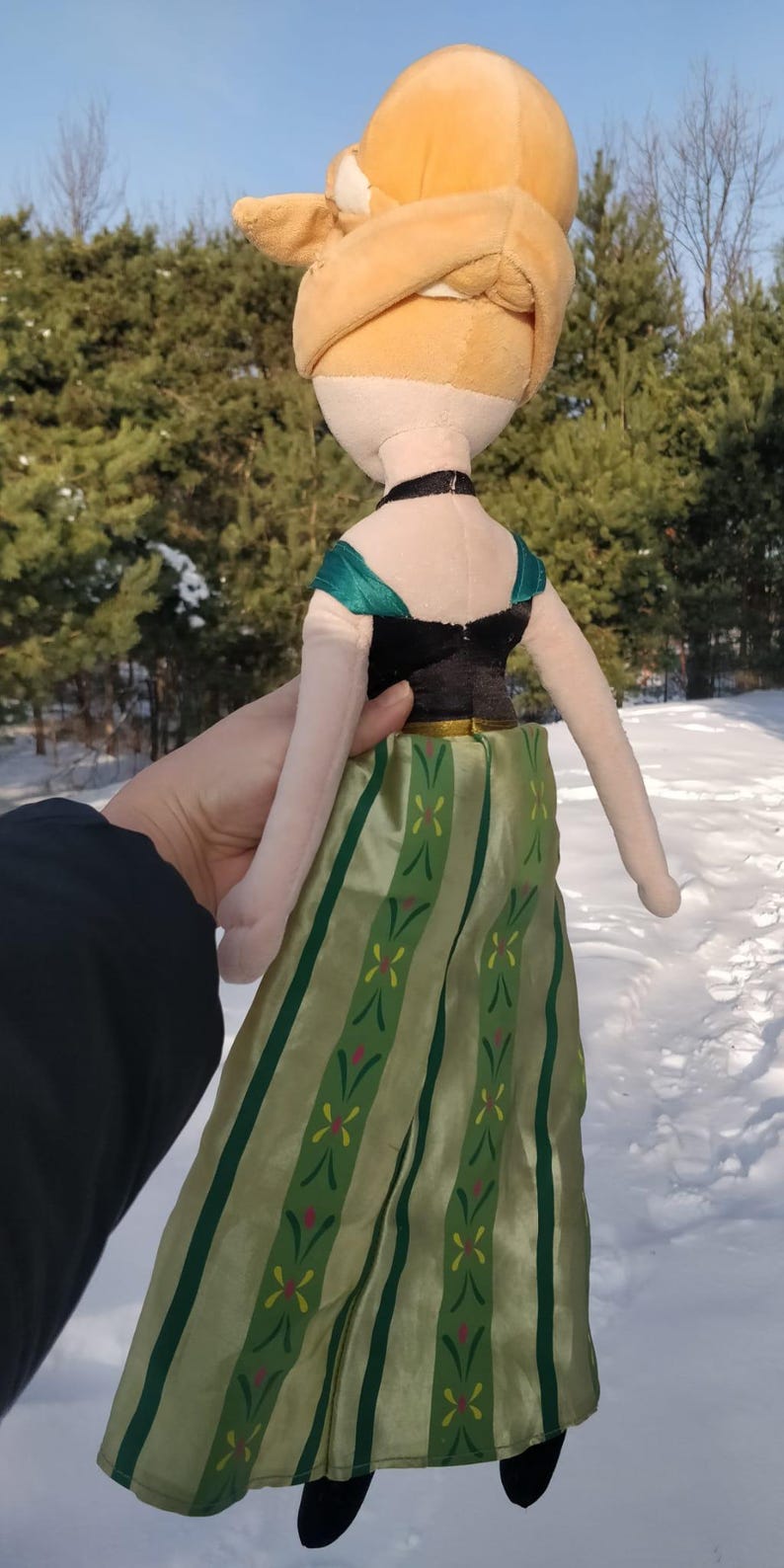 Op de afbeelding: Een pluche pop van een personage met blond haar in een knot. De pop draagt een zwarte top met groene accenten en een lange groene rok met een bloemenpatroon. De pop wordt vastgehouden tegen een besneeuwde achtergrond.