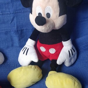 Puede incluir: Un juguete de peluche de Mickey Mouse con orejas negras, cuerpo negro, pantalones cortos rojos con dos botones blancos y zapatos amarillos. El juguete est&aacute; sentado sobre una superficie de tela azul.