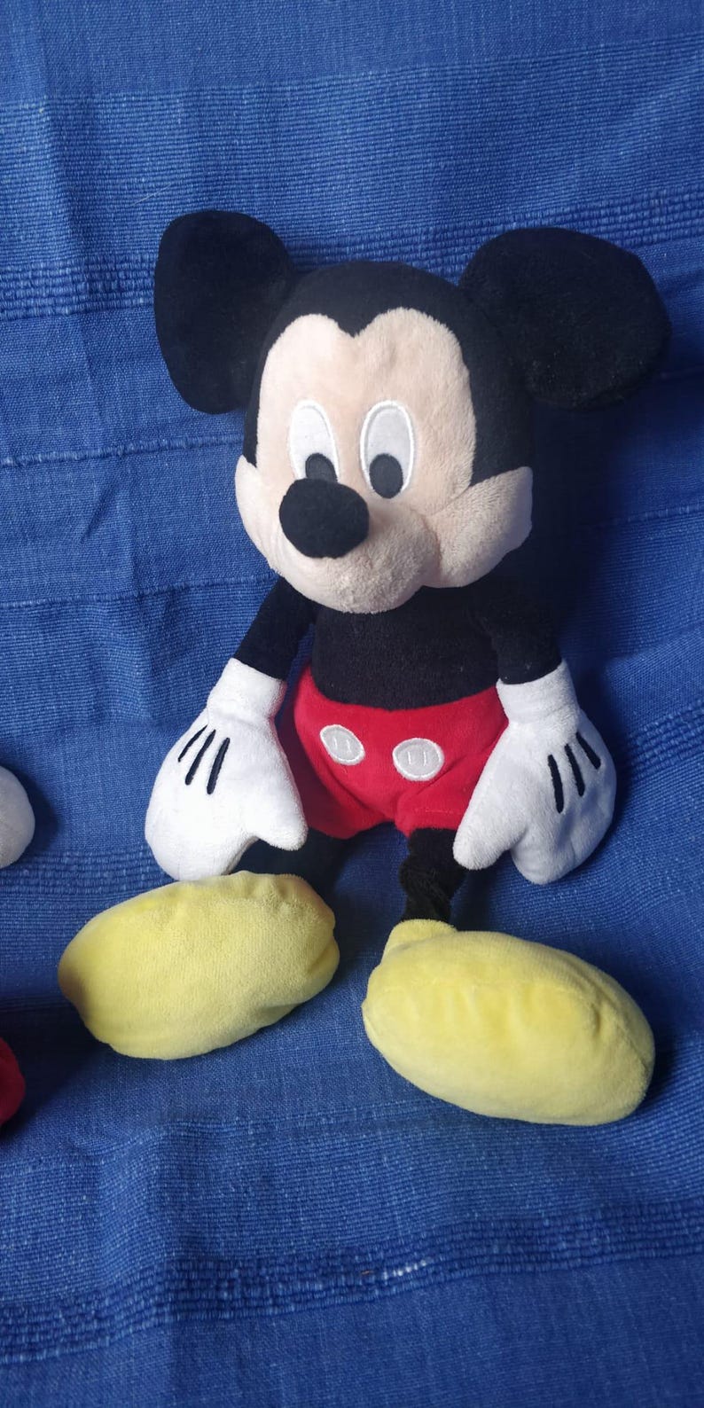 Puede incluir: Un juguete de peluche de Mickey Mouse con orejas negras, cuerpo negro, pantalones cortos rojos con dos botones blancos y zapatos amarillos. El juguete est&aacute; sentado sobre una superficie de tela azul.