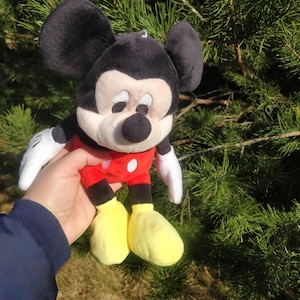Puede incluir: Un juguete de peluche de Mickey Mouse con orejas negras, nariz negra y zapatos amarillos. El juguete lleva una camisa roja con lunares blancos.