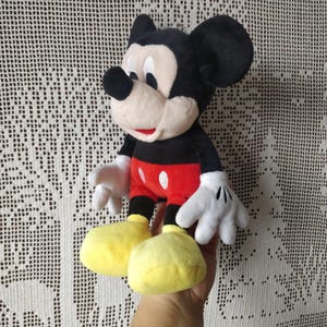 Puede incluir: Un juguete de peluche de Mickey Mouse con pelaje negro, pantalones cortos rojos y zapatos amarillos. El juguete tiene una gran sonrisa y orejas grandes y redondas.