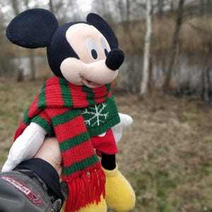 Peut inclure: Un jouet en peluche Mickey Mouse portant une &eacute;charpe ray&eacute;e rouge et verte avec un motif flocon de neige. Le jouet tient une mitaine rouge et blanche.