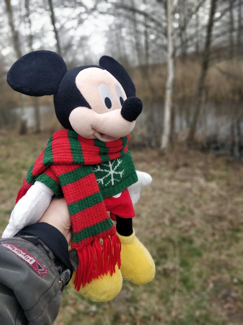 Peut inclure: Un jouet en peluche Mickey Mouse portant une &eacute;charpe ray&eacute;e rouge et verte avec un motif flocon de neige. Le jouet tient une mitaine rouge et blanche.