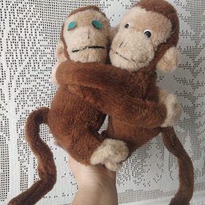 Può includere: Due scimmie di peluche marroni che si abbracciano. Le scimmie hanno facce beige, pelliccia marrone e lunghe code. Una scimmia ha gli occhi blu. Questi giocattoli morbidi e coccolosi sono perfetti per i bambini o come decorazione.